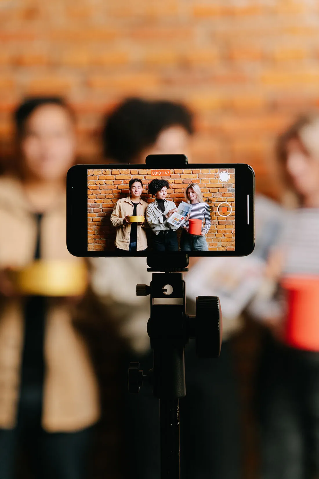 A phone on a tripod filming three people holding branded products against a brick wall during a content shoot.